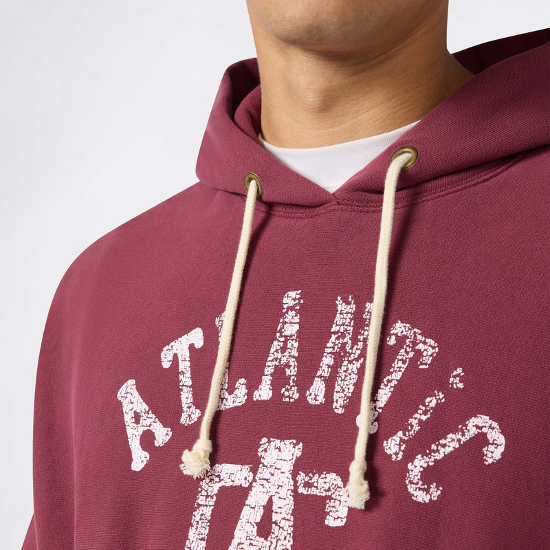A person wearing a maroon Champion Men’s Knitted Hooded Sweatshirt made from heavyweight fleece, featuring white drawstrings and a faded white “ATLANTIC” print on the front. The photo displays the hoodie from shoulders to mid-chest.