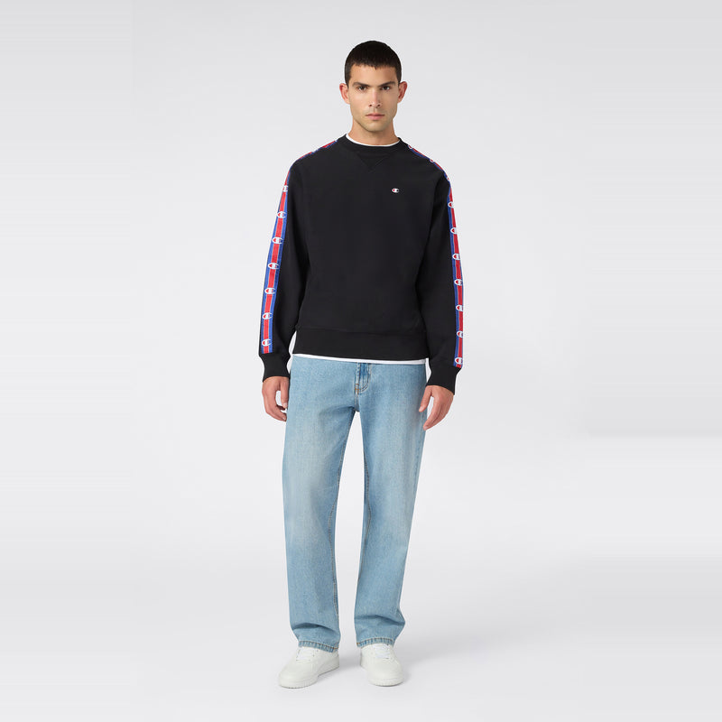 A person stands facing forward, wearing the Champion Men’s Knitted Crewneck Sweatshirt in black, featuring colorful logo stripes on the sleeves, paired with light blue jeans and white sneakers, against a plain light gray background. The cozy fleece and signature Reverse Weave construction of the sweatshirt add a classic touch to this relaxed look.
