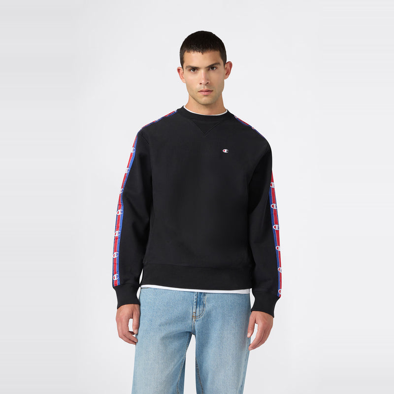 A man stands facing forward against a plain white background, wearing the Champion Men’s Knitted Crewneck Sweatshirt in black, featuring soft fleece fabric and logo tape on the sleeves, paired with light blue jeans.