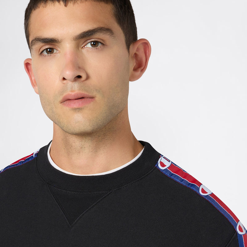 A young man with short brown hair is wearing the Champion Men’s Knitted Crewneck Sweatshirt, featuring cozy fleece and red and blue patterned trim on the shoulders, as he stands against a plain white background.