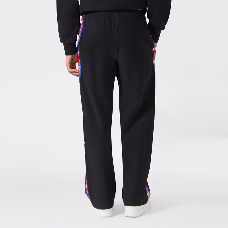 Back view of a person wearing black Champion Men’s Knitted Straight Hem Pants featuring red, white, and blue logo tape along the sides, paired with a matching black sweatshirt and white sneakers against a plain light gray background—an ideal option for men’s sportswear.