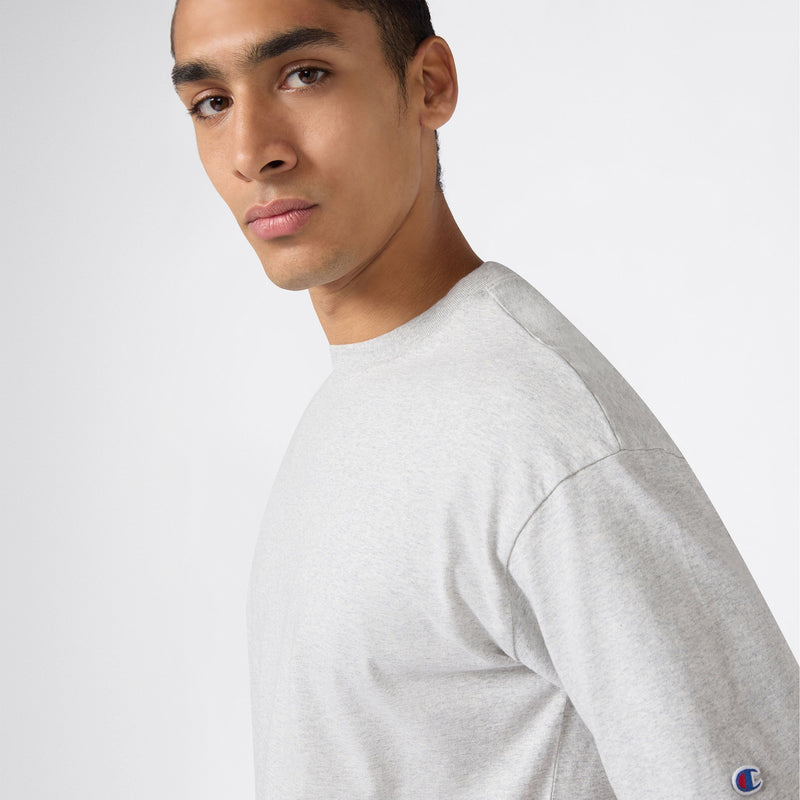 A man wearing a light gray Champion Men’s Knitted T-Shape Tee faces the camera against a plain white background, highlighting this classic piece of men’s sportswear.