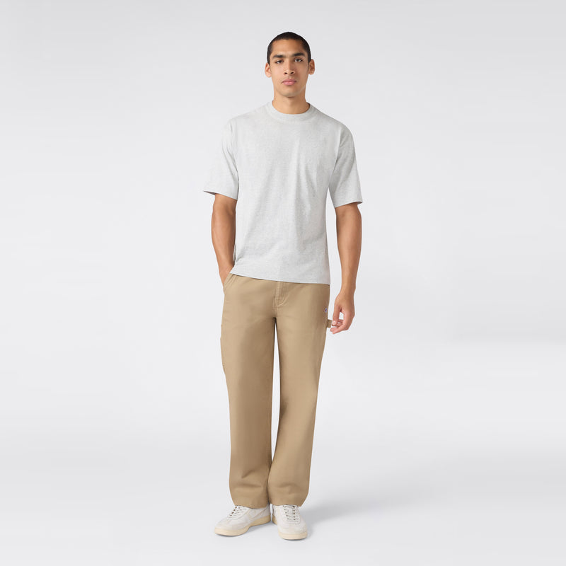 A person stands facing forward, wearing the Champion Men’s Knitted T-Shape Tee in light gray, paired with tan pants and white sneakers against a plain light background—a modern take on men’s sportswear.