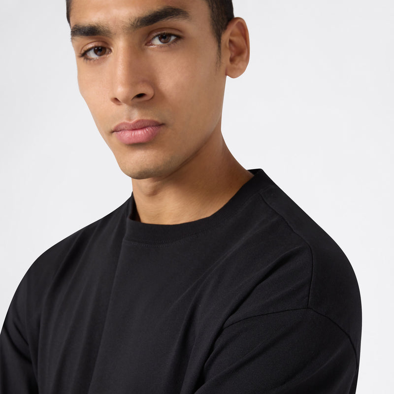 A person with short dark hair wearing a black Champion Men’s Knitted T-Shape Tee looks seriously at the camera against a plain white background.