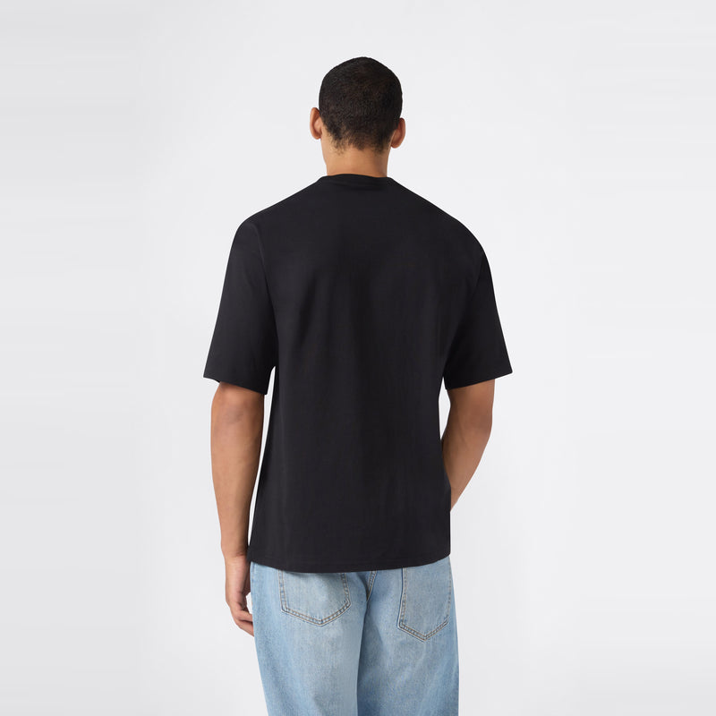 A person stands facing away from the camera, wearing a plain black Champion Men’s Knitted T-Shape Tee and light blue jeans against a neutral, light background.
