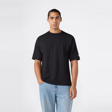 A person wearing a Champion Men’s Knitted T-Shape Tee in plain black and light blue jeans stands against a simple, light gray background, with one hand in their pocket, facing forward.
