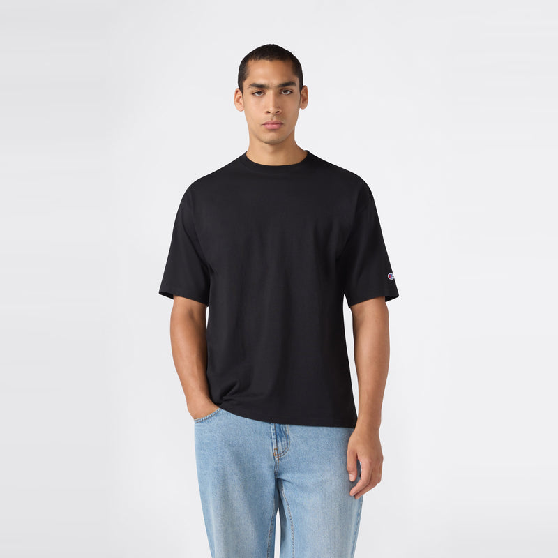 A person wearing a Champion Men’s Knitted T-Shape Tee in plain black and light blue jeans stands against a simple, light gray background, with one hand in their pocket, facing forward.