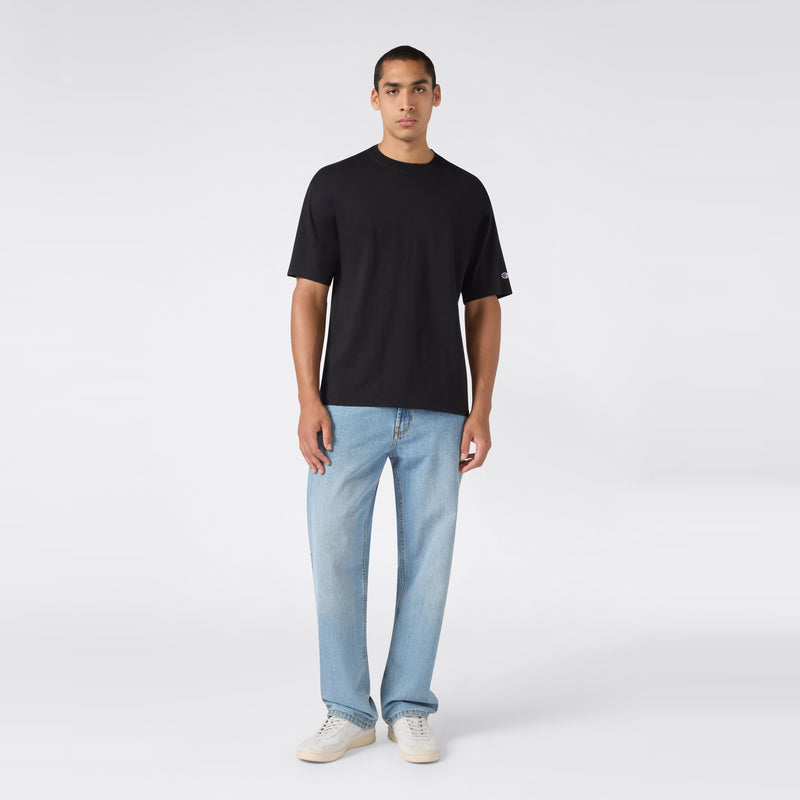 A person stands facing forward, wearing a plain black Champion Men’s Knitted T-Shape Tee, light blue jeans, and white sneakers, against a plain white background.
