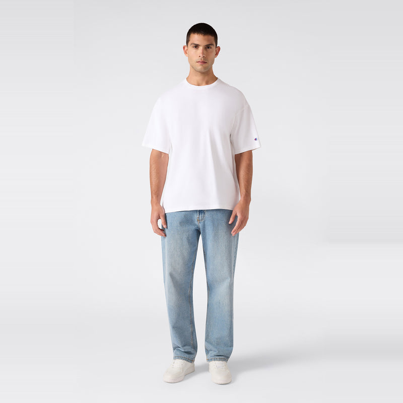 A person stands facing forward, wearing the Champion Men’s Knitted T-Shape Tee, paired with light blue jeans and white sneakers, against a plain light gray background.