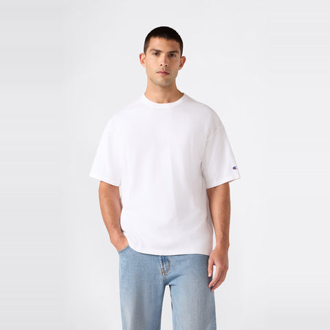 A man wearing the Champion Men’s Knitted T-Shape Tee in plain white and light blue jeans stands against a plain, light gray background, looking directly at the camera with one hand in his pocket.