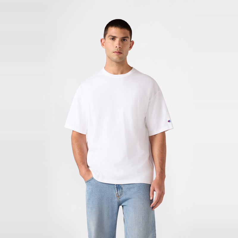 A man wearing the Champion Men’s Knitted T-Shape Tee in plain white and light blue jeans stands against a plain, light gray background, looking directly at the camera with one hand in his pocket.