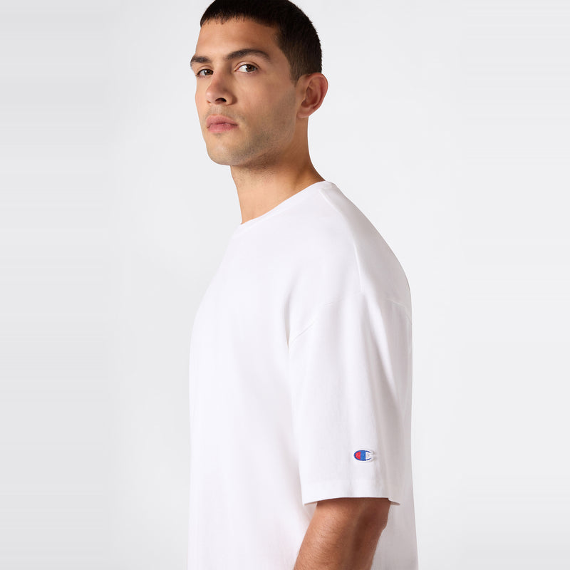 A man with short dark hair stands against a light gray background, looking slightly to the left. He’s wearing the Champion Men’s Knitted T-Shape Tee—a plain white, heavyweight cotton t-shirt featuring a small Champion logo on the left sleeve—offering a sleek example of timeless men’s sportswear.