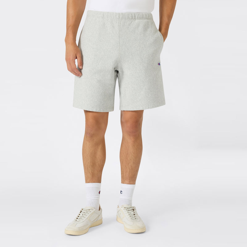 A person wearing Men’s Knitted Shorts by Champion in light gray, paired with white socks and cream sneakers, stands against a plain background. Their hands rest in their pockets, and only the lower half of their body is visible—showcasing casual men’s sportswear from Champion crafted from heavyweight fleece.