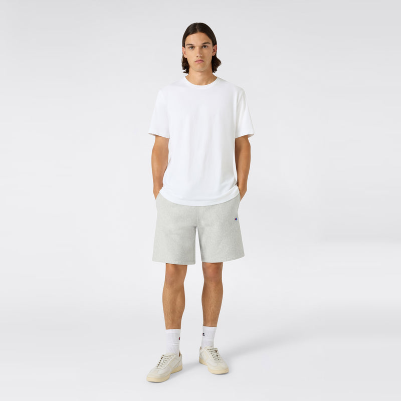 A person with long hair stands facing forward, wearing a plain white t-shirt and Champion Men’s Knitted Shorts in light gray—a staple of men’s sportswear—along with white socks and white sneakers, against a simple light gray background. Their hands are in their pockets.