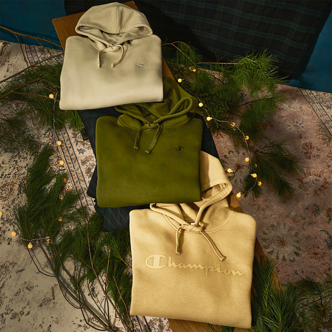 Three folded hoodies in beige, green, and brown on a textured surface with decorative lights and plants.