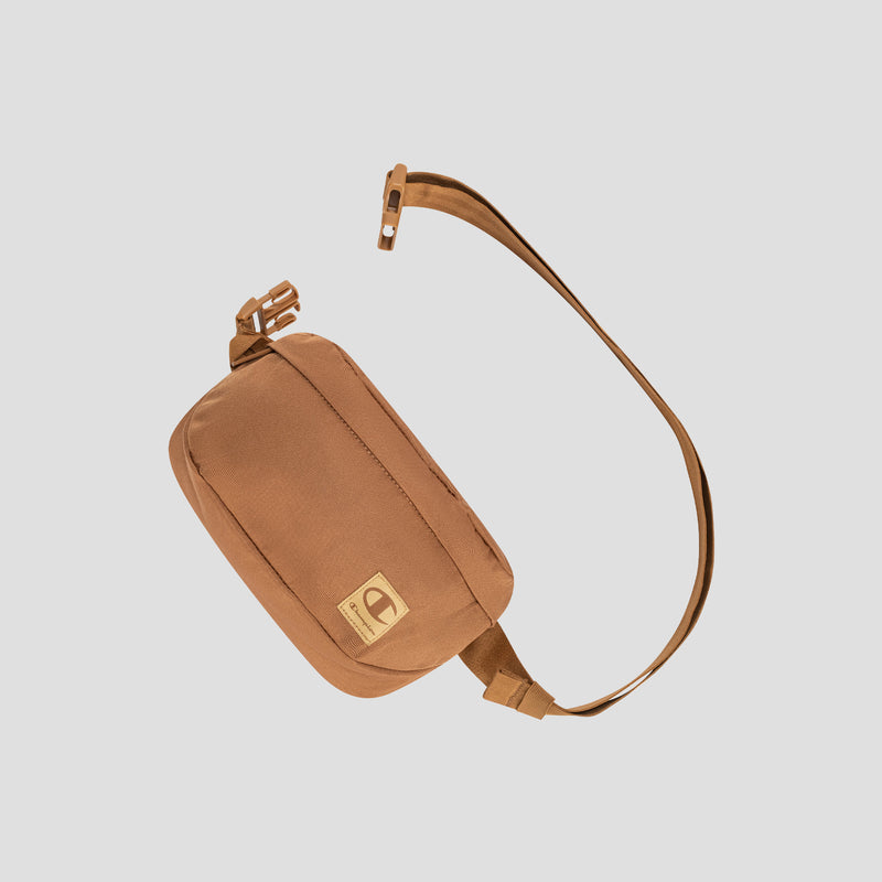 A compact, rectangular tan Atlas Waist Pack by Champion with a long adjustable strap for hands-free convenience and a front patch featuring the Champion logo, displayed on a light gray background.