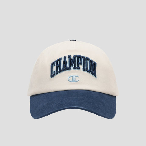 A beige and navy blue Amor Adjustable Baseball Cap by Champion, crafted from washed cotton twill and featuring “CHAMPION” embroidered in dark blue lettering on the front with a small Champion logo below, photographed on a plain gray background—showcasing true Champion authenticity.