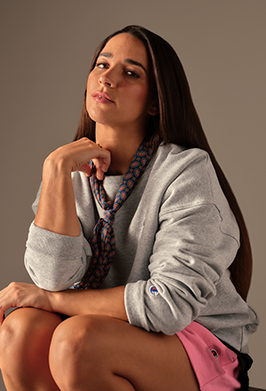 Woman wearing a gray sweater and pink shorts sitting against a plain background