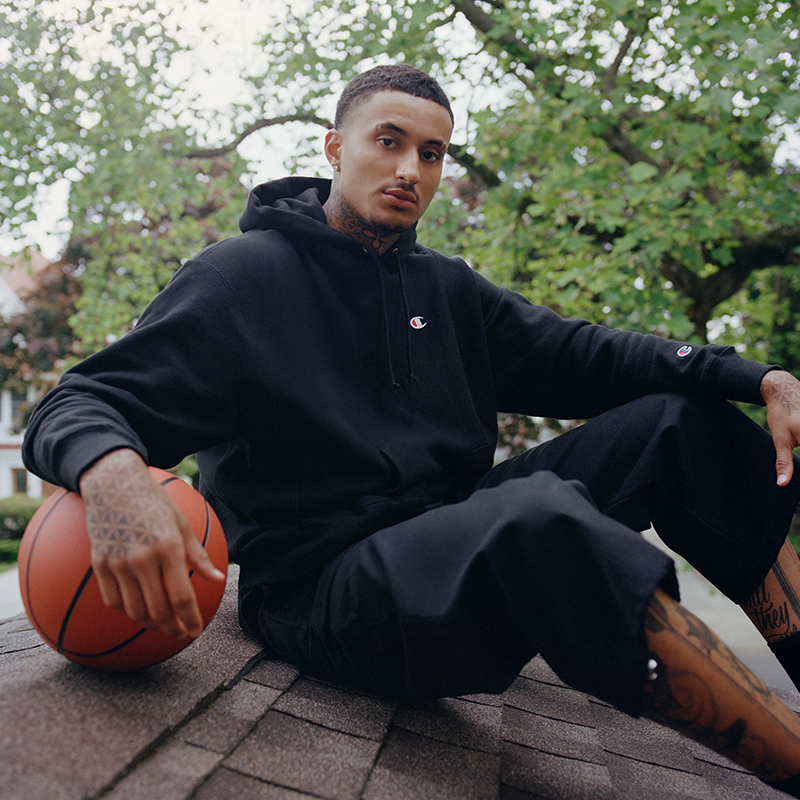 A man wearing a black Champion hoodie and matching black sweatpants sits on a roof holding a basketball, with trees and a house in the background.