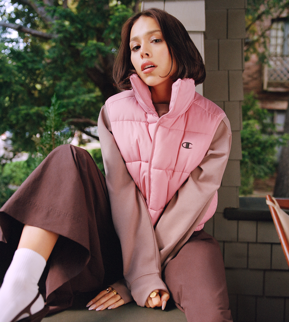 A woman wearing a pink puffer vest over a dusty pink long-sleeve top and brown wide-leg pants, sitting outdoors on a porch with green foliage and a brick wall in the background. A small Champion 'C' logo is visible on the chest of the vest.