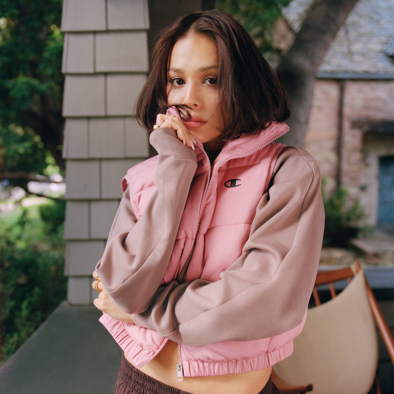 A woman wearing a pink puffer vest over a dusty mauve-brown cropped sweatshirt, standing on a porch with a house and outdoor greenery in the background.