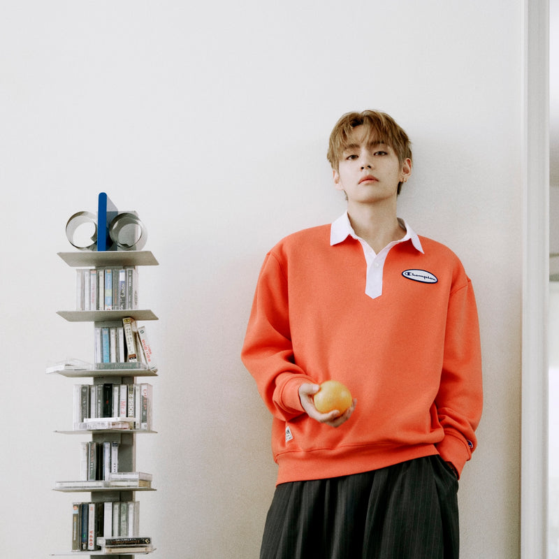 A person wearing a Champion Fleece Rugby stands next to a tall, stacked CD rack against a white wall while holding an orange.