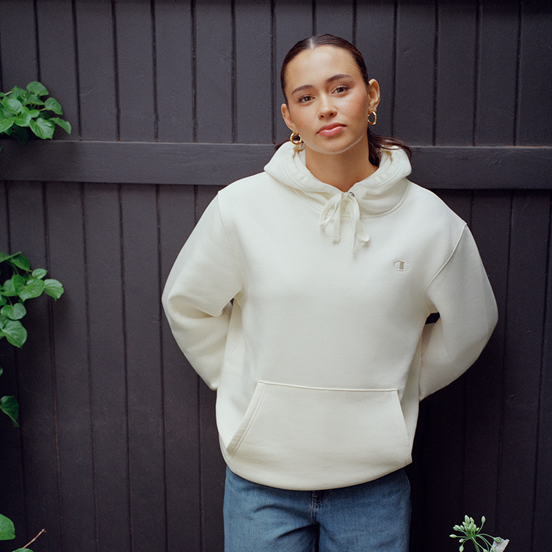 A woman wearing a cream-colored pullover hoodie with drawstrings and a kangaroo pocket, paired with blue jeans, standing against a dark gray wooden fence.