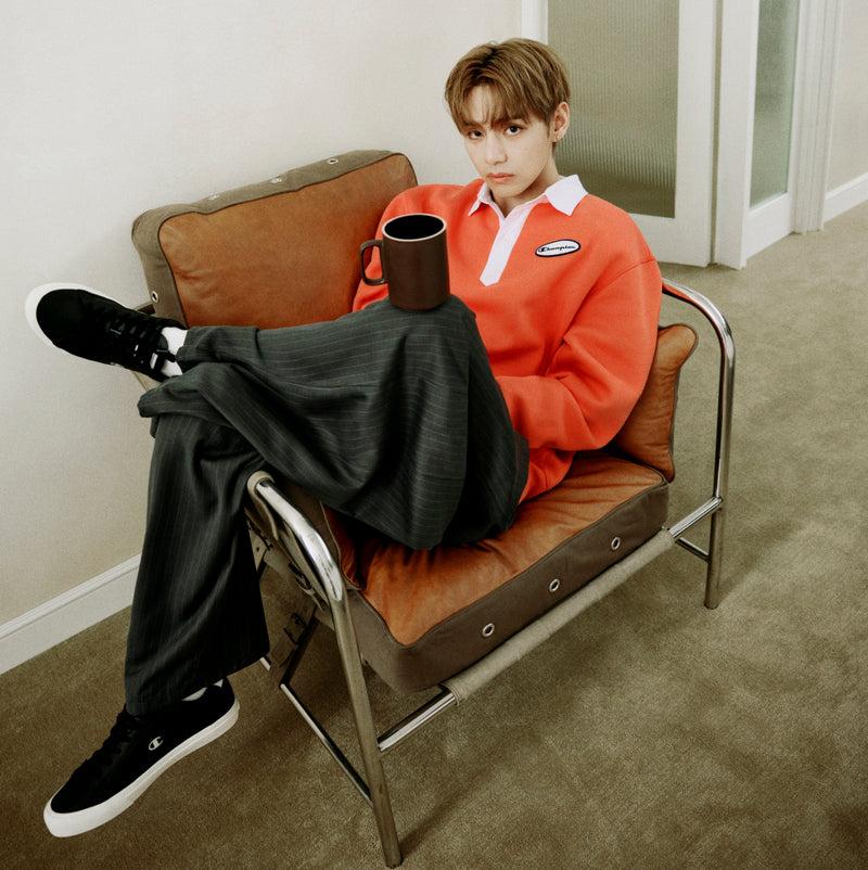 A person wearing a red Champion Fleece Rugby shirt sits cross-legged on a brown chair, holding a mug and looking at the camera. They pair the shirt with dark pants and black sneakers, set against the backdrop of a neutral-colored room.