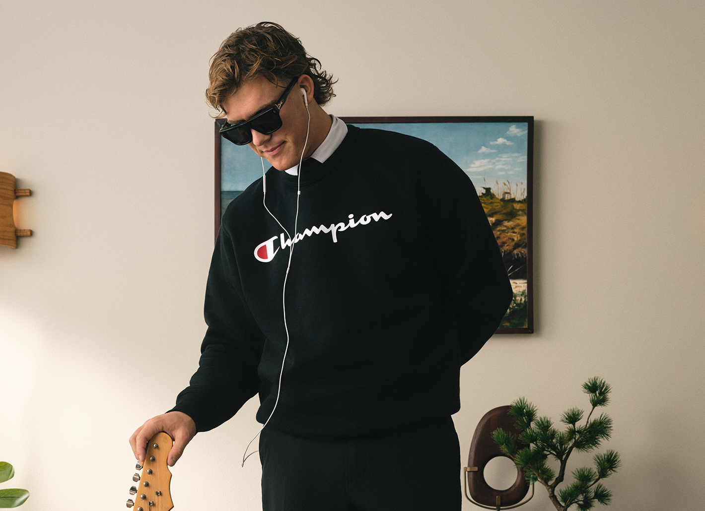 Person wearing a black Champion sweatshirt with sunglasses, standing in a room with a painting on the wall.