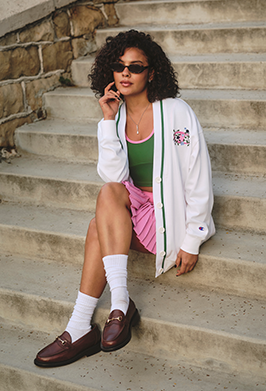 Woman sitting on stone steps wearing a white cardigan with a pink design, green top, pink skirt, and brown shoes.