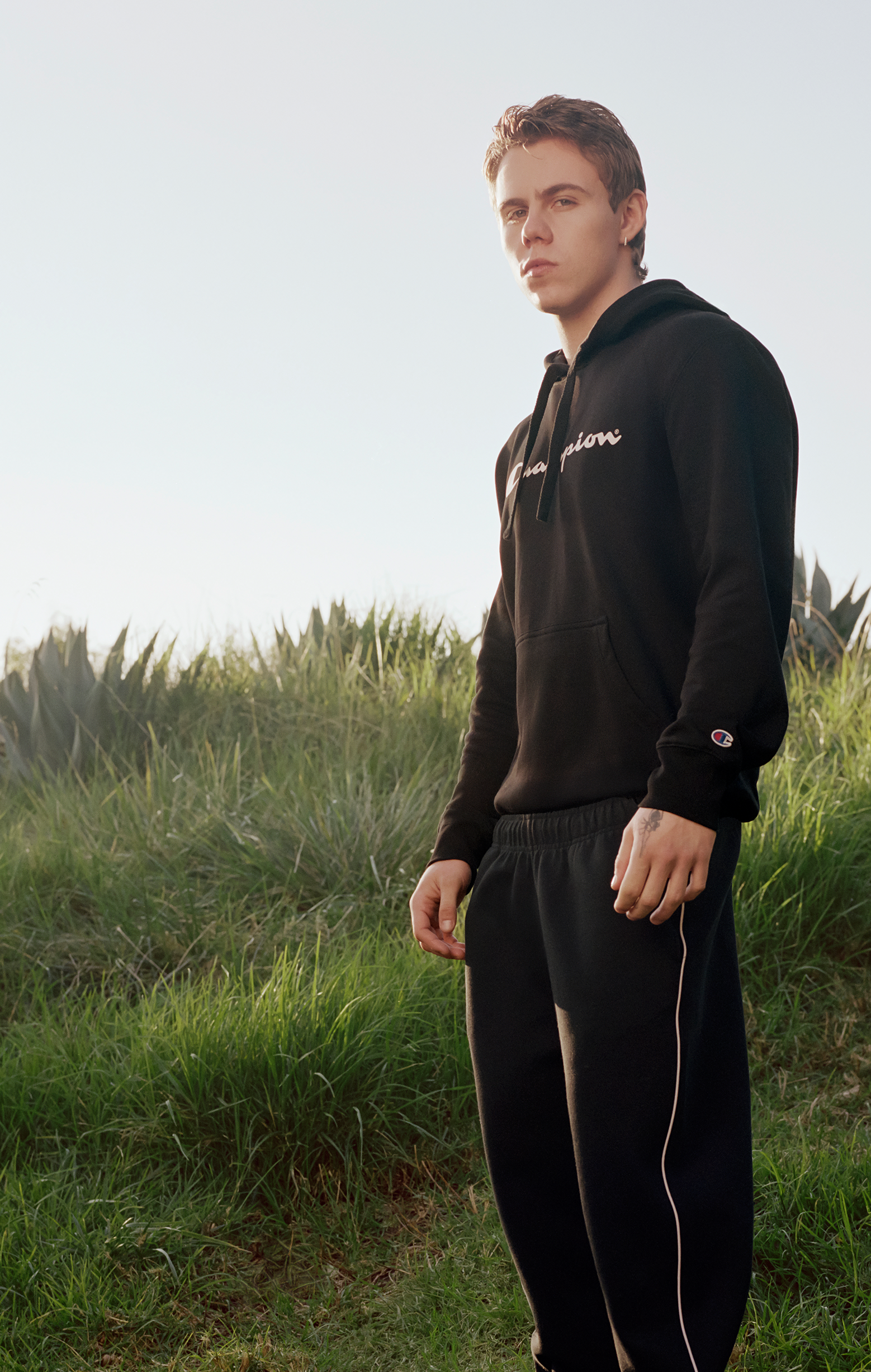 A person stands outdoors on grass wearing a black Champion hoodie and matching black pants with white piping, looking towards the camera with a neutral expression. Tall green plants and a clear sky are in the background.