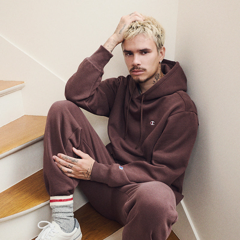A man with blonde hair sits on wooden stairs wearing a matching brown Champion hoodie and sweatpants set, white sneakers, and gray socks with red stripes.