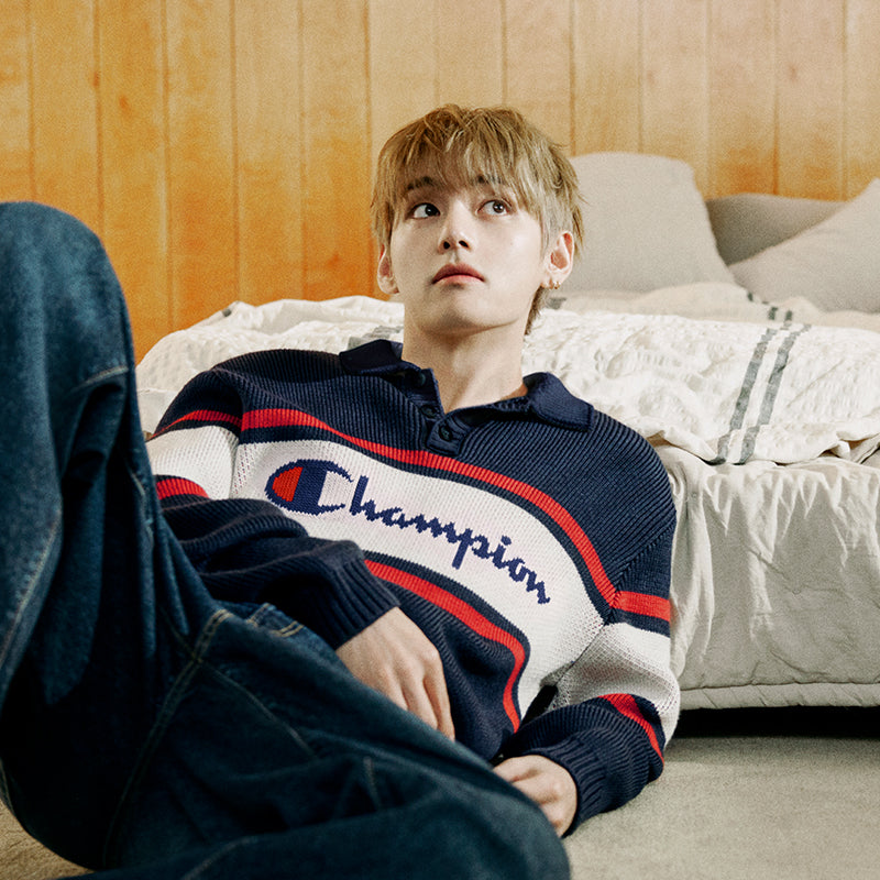 A man reclining on a bed wearing a navy blue Champion knit polo sweater with red and white stripes and blue jeans, set against a warm wood-paneled bedroom background.