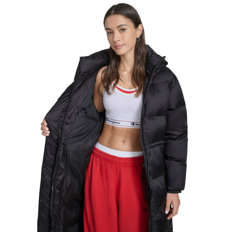A woman wearing the Champion Maxi Length Cinch Waist Hooded Puffer over a white sports bra and red pants stands against a white background, holding one side of the coat open to display her outfit.