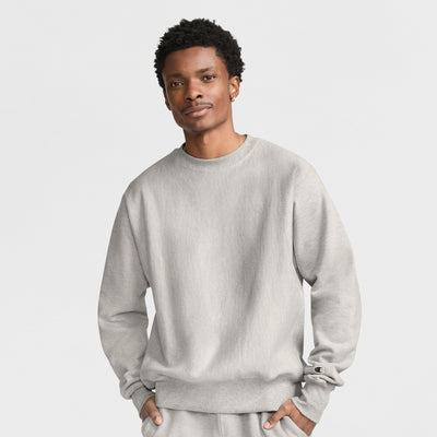 A man wears a light gray Champion Reverse Weave Crewneck Sweatshirt with matching pants, hands in pockets, looking at the camera against a plain white background.