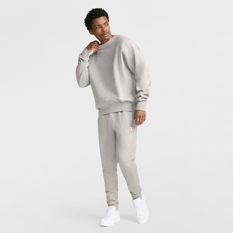 A person stands against a plain white background, wearing a light gray Champion Reverse Weave Crewneck Sweatshirt and matching sweatpants with white sneakers, looking to the side while casually touching their neck.