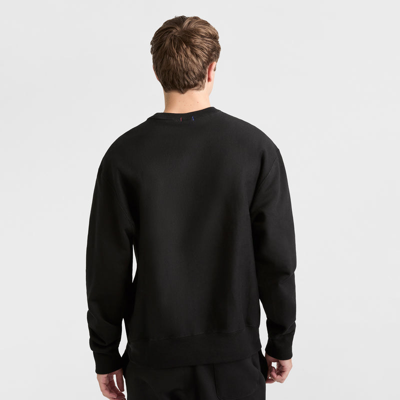 A person with short brown hair stands facing away from the camera, wearing a plain black Champion Reverse Weave Crewneck Sweatshirt and black pants against a white background.