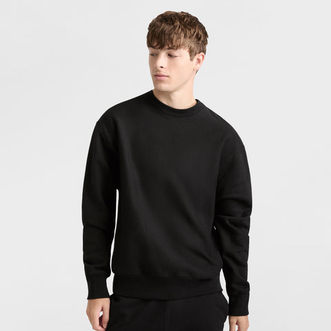 A young person with short brown hair models the Champion Reverse Weave Crewneck Sweatshirt in black with matching pants, standing against a plain white background and gazing slightly to the side.