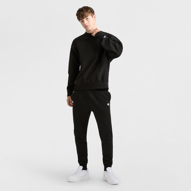 A person stands against a plain white background wearing a black Champion Reverse Weave Crewneck Sweatshirt and black jogger pants with white sneakers, posing with one hand touching their neck.
