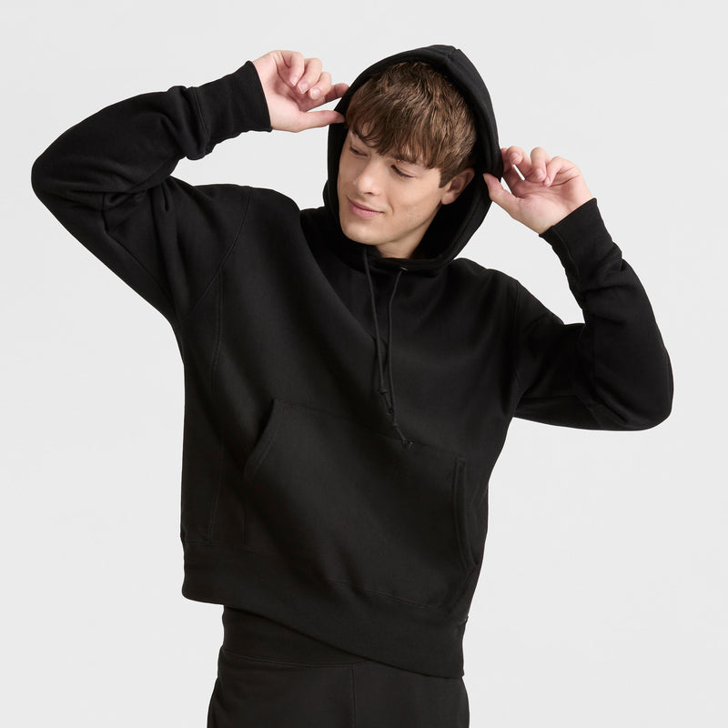 A person in a black Champion Reverse Weave Hoodie and black pants stands against a plain light background, holding the hood with both hands and looking down with a slight smile.