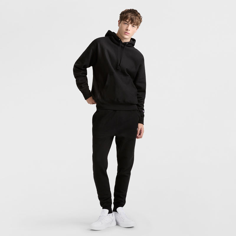 A young man with short brown hair and a relaxed posture stands against a white background, wearing a Champion Reverse Weave Hoodie in black, black jogger pants, and white sneakers, with one hand in his pocket.