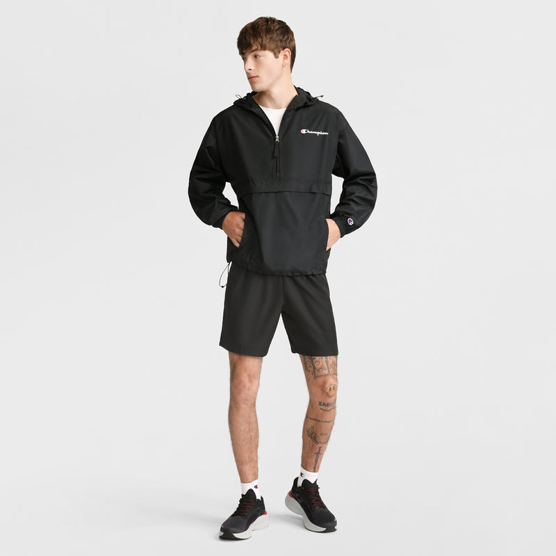 A person stands against a white background, wearing a black Champion jacket, Champion 7" Woven Sport Shorts, white socks, and black sneakers. They have tattoos on one leg and look to the side with hands in their jacket pockets.