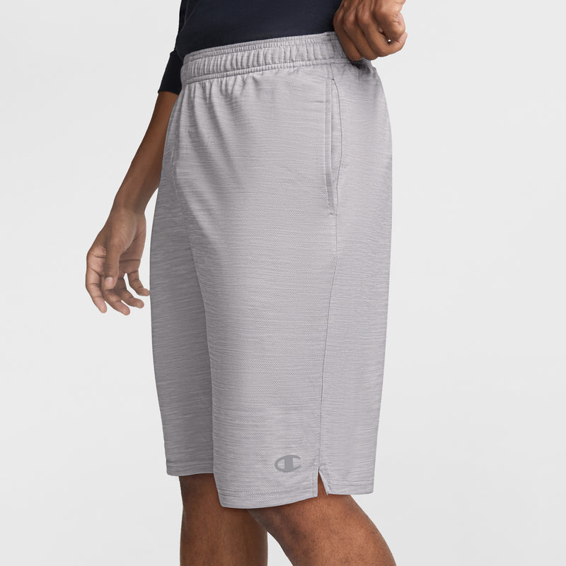 A man wears Champion 10" Moisture Wicking Sport Shorts in light gray with an elastic waistband, side pockets, and logo near the hem, standing against a white background. These athletic shorts offer comfort and moisture control.