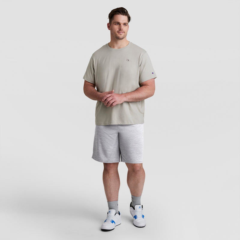 A man stands against a plain light background wearing light gray Champion 10" Moisture Wicking, Sport Shorts - Big & Tall, a matching t-shirt, gray socks, and white sneakers. He loosely clasps his hands in front and looks slightly to the side.