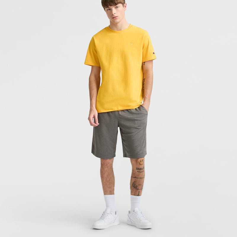 A young man with short hair and tattooed legs stands against a white background, wearing a yellow t-shirt, Champion 10" Moisture Wicking Sport Shorts, white socks, and sneakers, with one hand in his pocket.