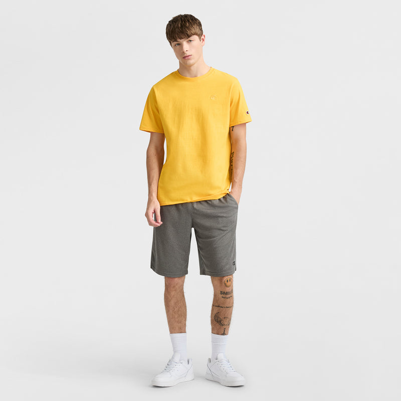 A young man with short brown hair and tattoos on his left leg stands against a white background, wearing Champion 10" Moisture Wicking Sport Shorts, a yellow T-shirt, white socks, and white sneakers.