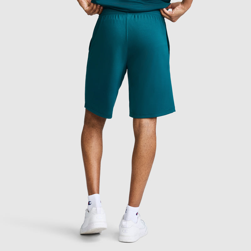 Standing with hands on hips, the person is seen from the back against a plain light background, wearing Champion 10″ Moisture Wicking Sport Shorts in teal, paired with white socks and white sneakers.
