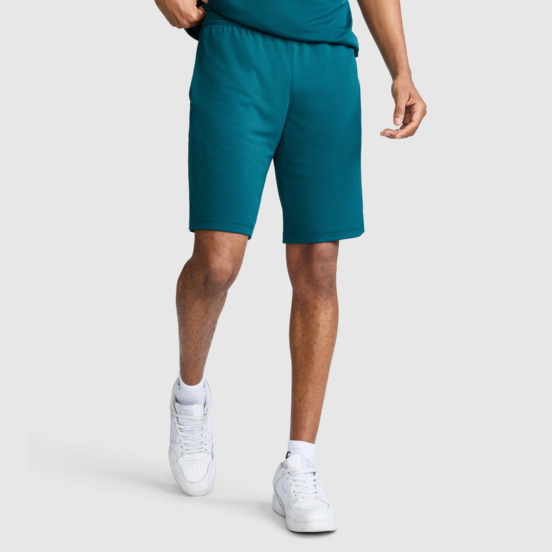 A person wearing Champion 10″ Moisture Wicking Sport Shorts in teal, a matching teal shirt, white socks, and white sneakers stands against a plain light gray background. Their face is not visible.