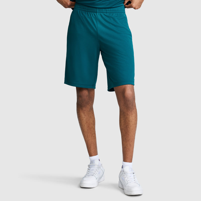 A person wearing Champion 10″ Moisture Wicking Sport Shorts in teal, paired with a matching shirt, white socks, and white sneakers, stands against a plain light gray background with hands on hips.