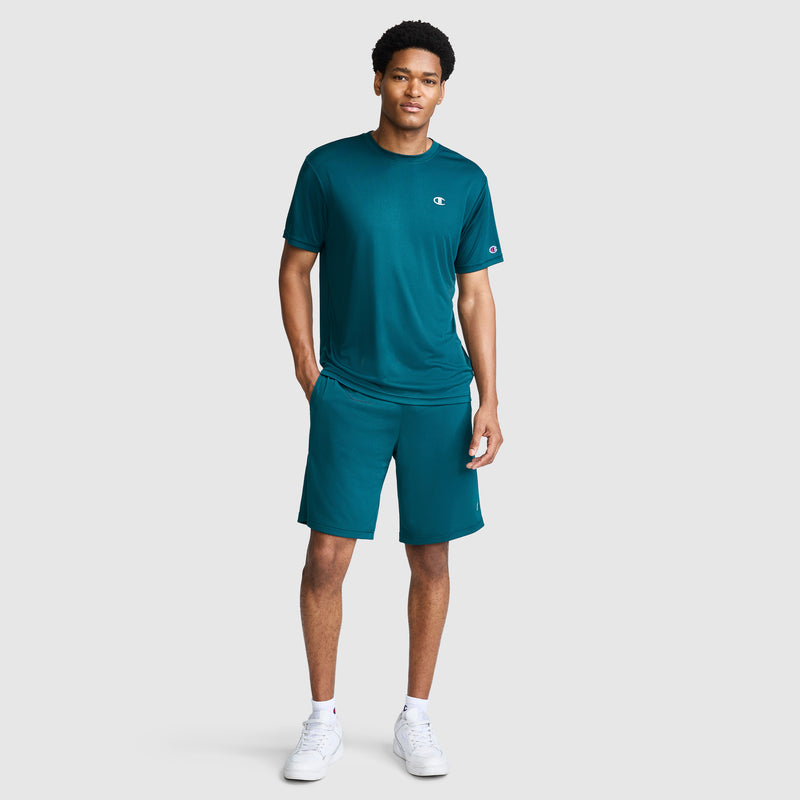 A man wearing a teal athletic t-shirt and Champion 10″ Moisture Wicking Sport Shorts stands with one hand in his pocket, looking forward. He is also wearing white sneakers against a plain, light gray background.